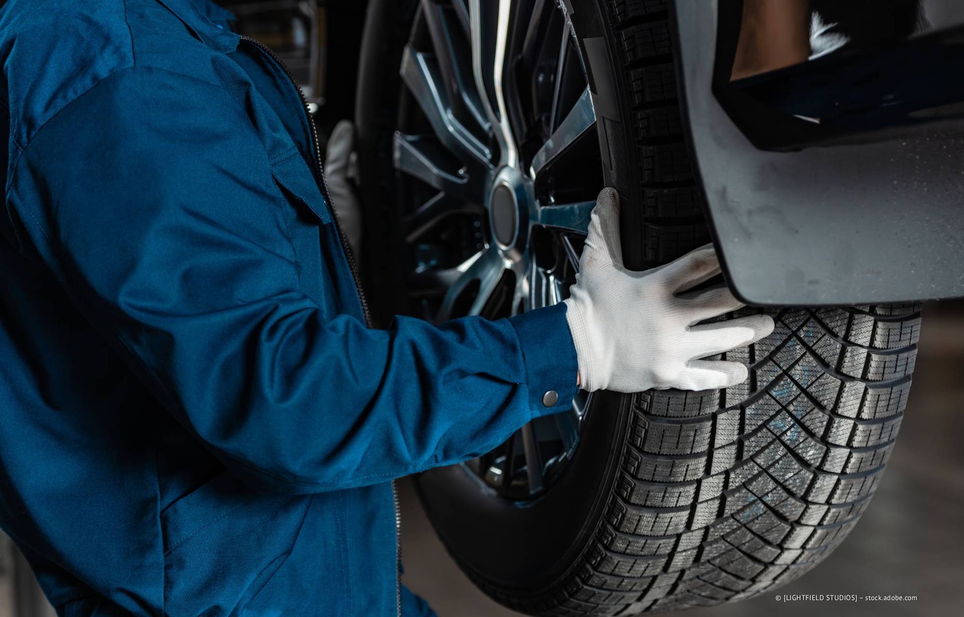 Mechaniker im blauen Overall, mit Handschuhen, hält einen Autoreifen.