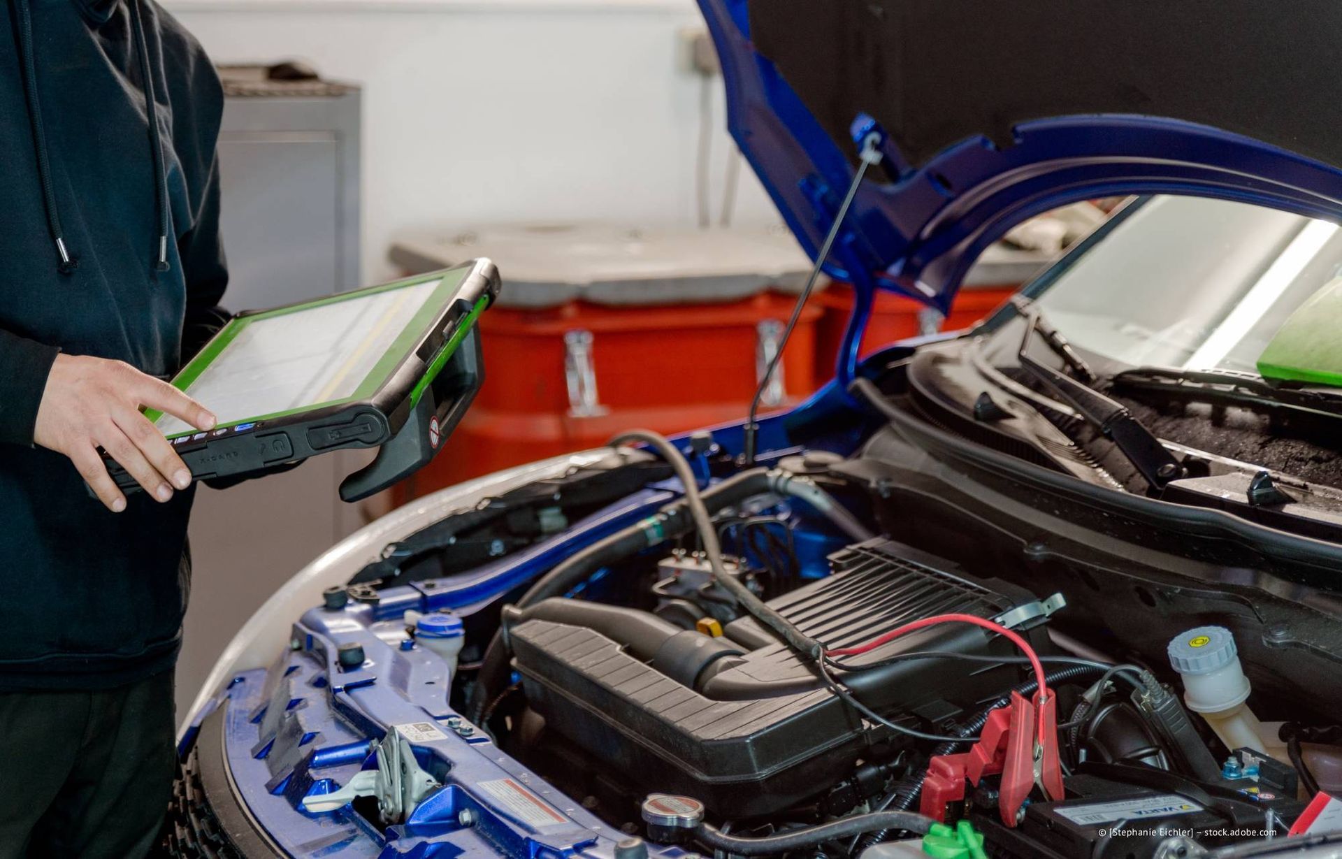 Ein Mechaniker benutzt ein Diagnose-Tablet, um in einer Werkstatt den Motor eines blauen Autos zu untersuchen.