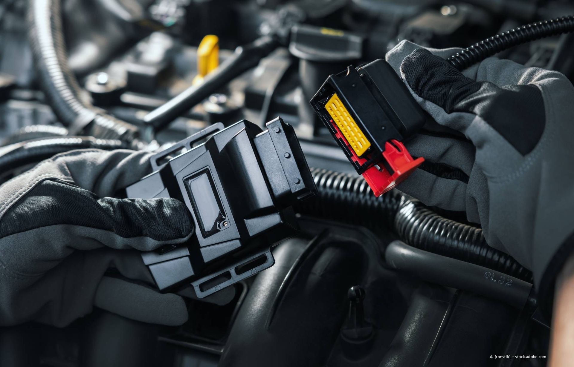 Hände in Handschuhen verbinden elektrische Steckverbinder im Motorraum eines Autos.