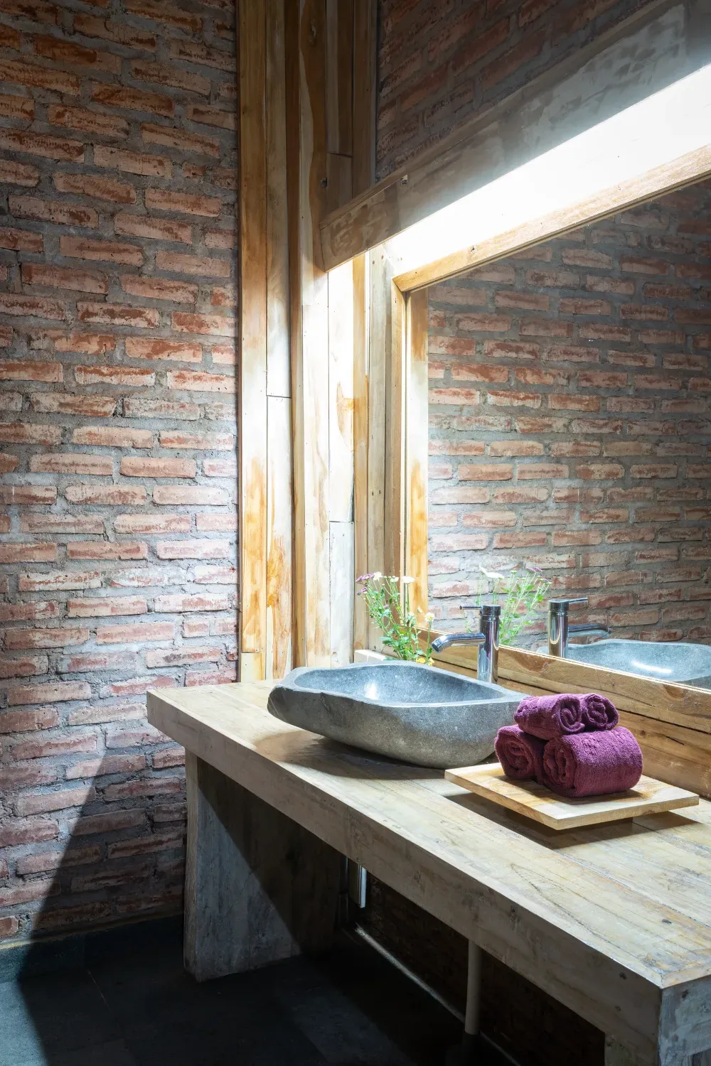 Baño con pared de ladrillo, tocador de madera, lavabo de piedra y toallas moradas.