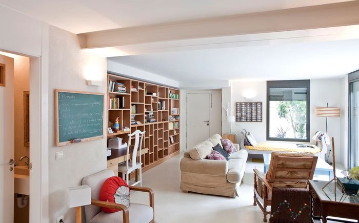 Luminoso salón con estanterías, pizarra, sofá beige y luz natural que entra por los grandes ventanales.