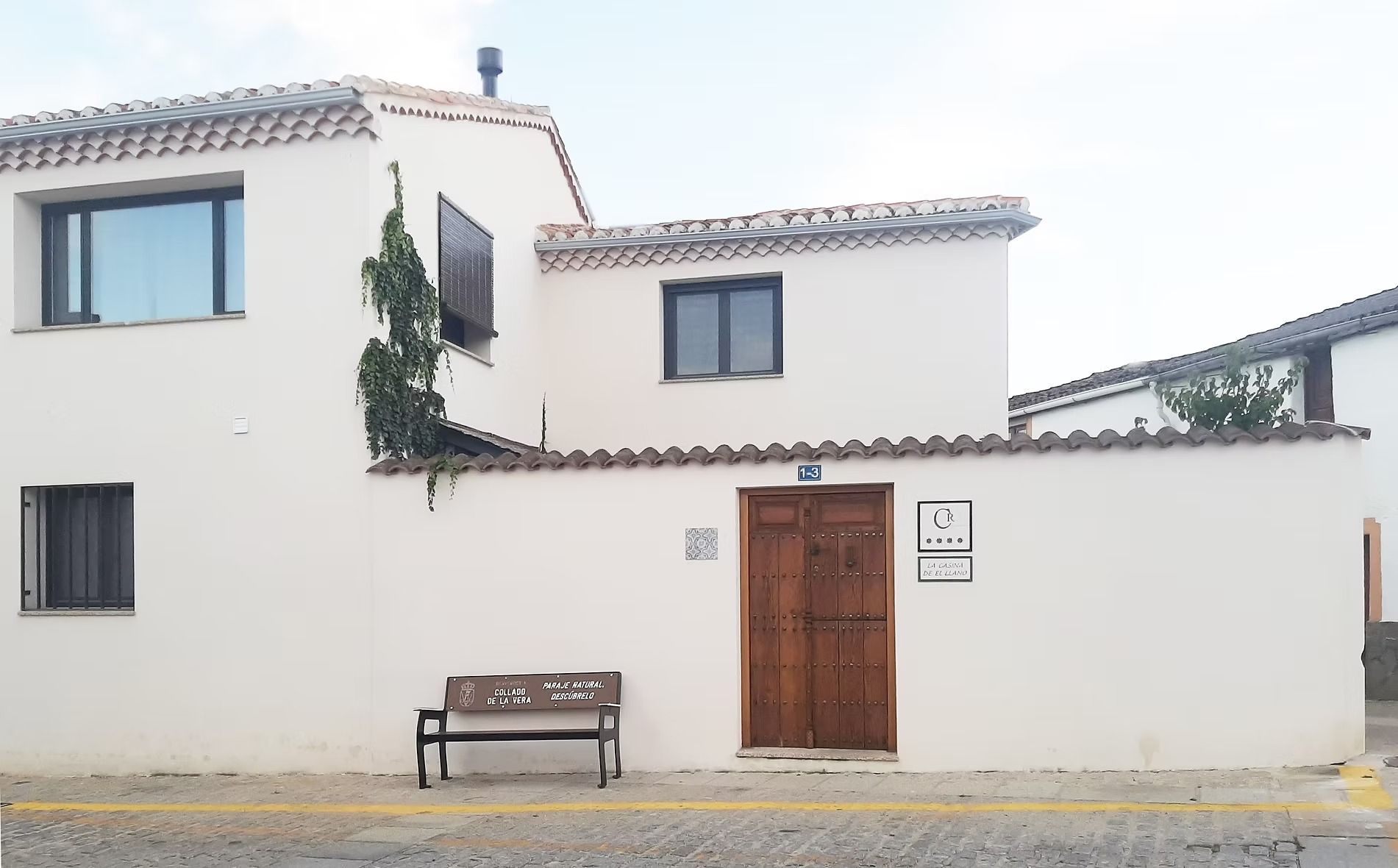 Casa de estuco blanco con ventanas oscuras, puerta de madera y un banco junto a la pared en una calle tranquila.