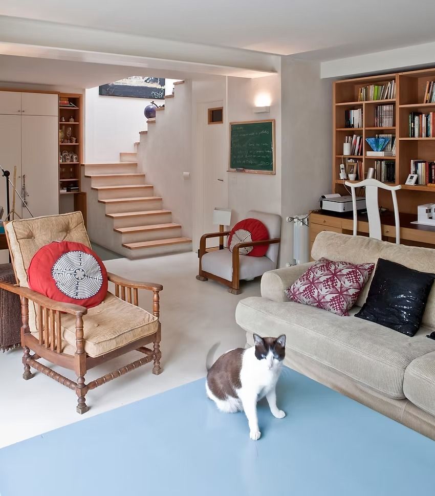 Luminoso salón con escalera, sillas de mimbre, sofá y un gato de pie sobre una mesa azul.