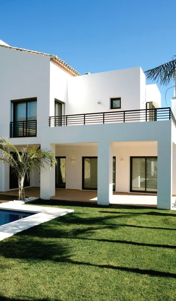 Moderna casa blanca de dos plantas con balcón, grandes ventanales y un césped verde bajo un cielo azul despejado.