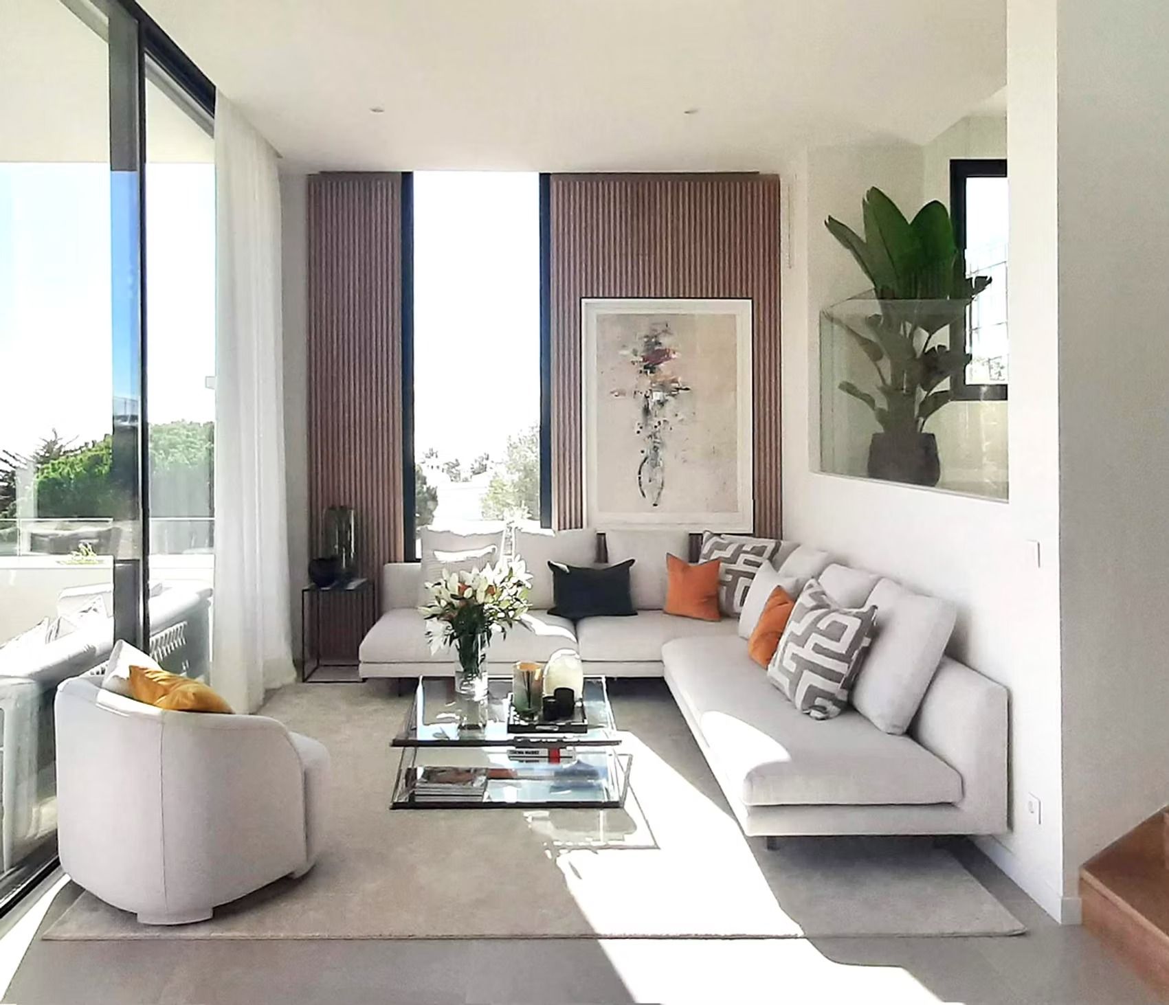Salón luminoso y moderno con sofá blanco, mesa de centro de cristal, plantas y grandes ventanales.