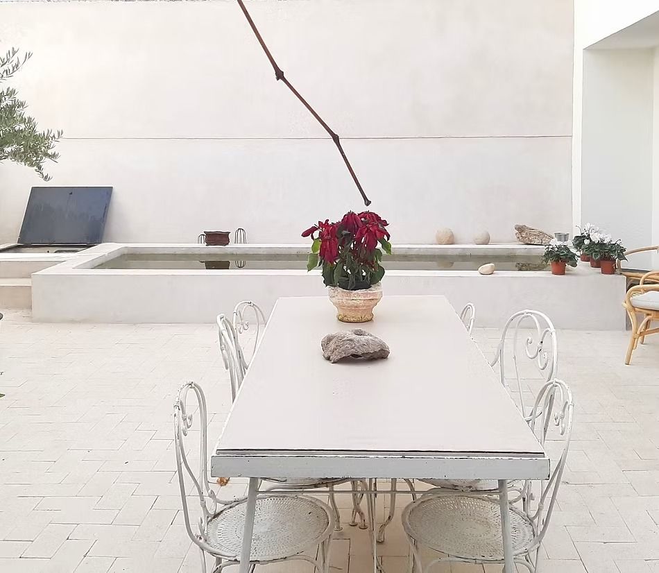 Mesa de comedor blanca para patio con sillas de mimbre, flores rojas y zona de estar exterior minimalista.