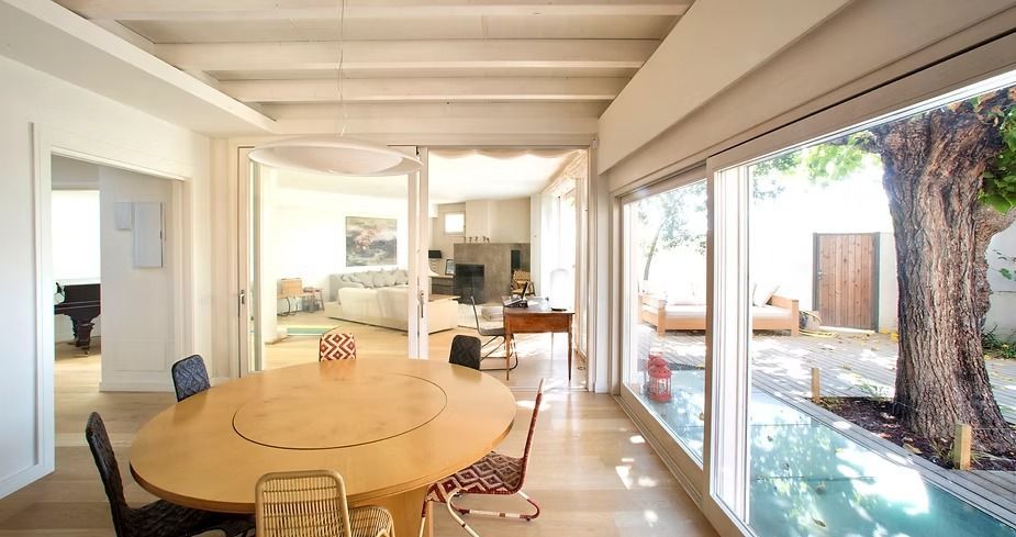 Comedor luminoso con mesa redonda de madera, sillas y ventanales que van del suelo al techo y dan a un patio con un árbol.