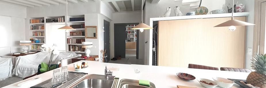 Cocina moderna con gabinetes blancos, fregadero de acero inoxidable, estantes abiertos y un luminoso comedor al fondo.