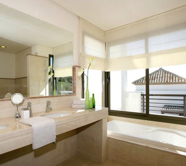 Baño moderno y luminoso con tocador de color beige, espejo grande y bañera junto a una ventana soleada.