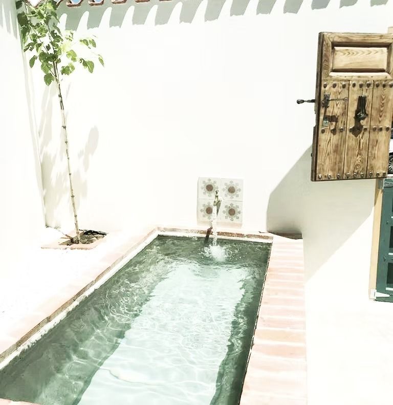 Patio estrecho con paredes blancas, piscina, pequeña fuente y armario rústico con puerta de madera.