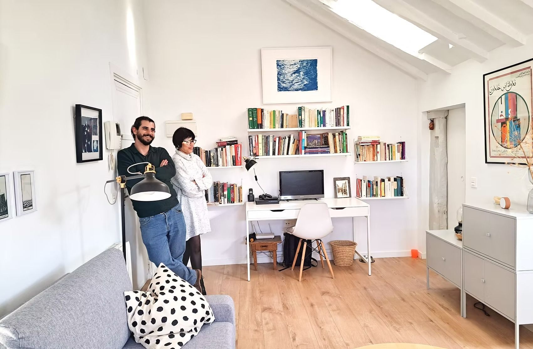 Despacho luminoso en casa con dos personas de pie junto a un escritorio, estanterías y un sofá en una habitación soleada.