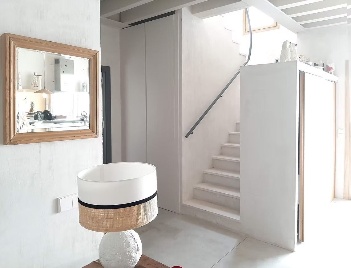 Pasillo luminoso con escalera, paredes blancas, espejo y mesa redonda en un interior minimalista.