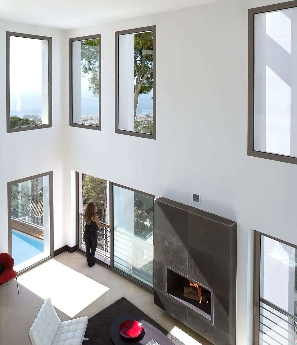 Luminoso salón moderno con grandes ventanales, chimenea y una persona junto a las puertas de cristal.