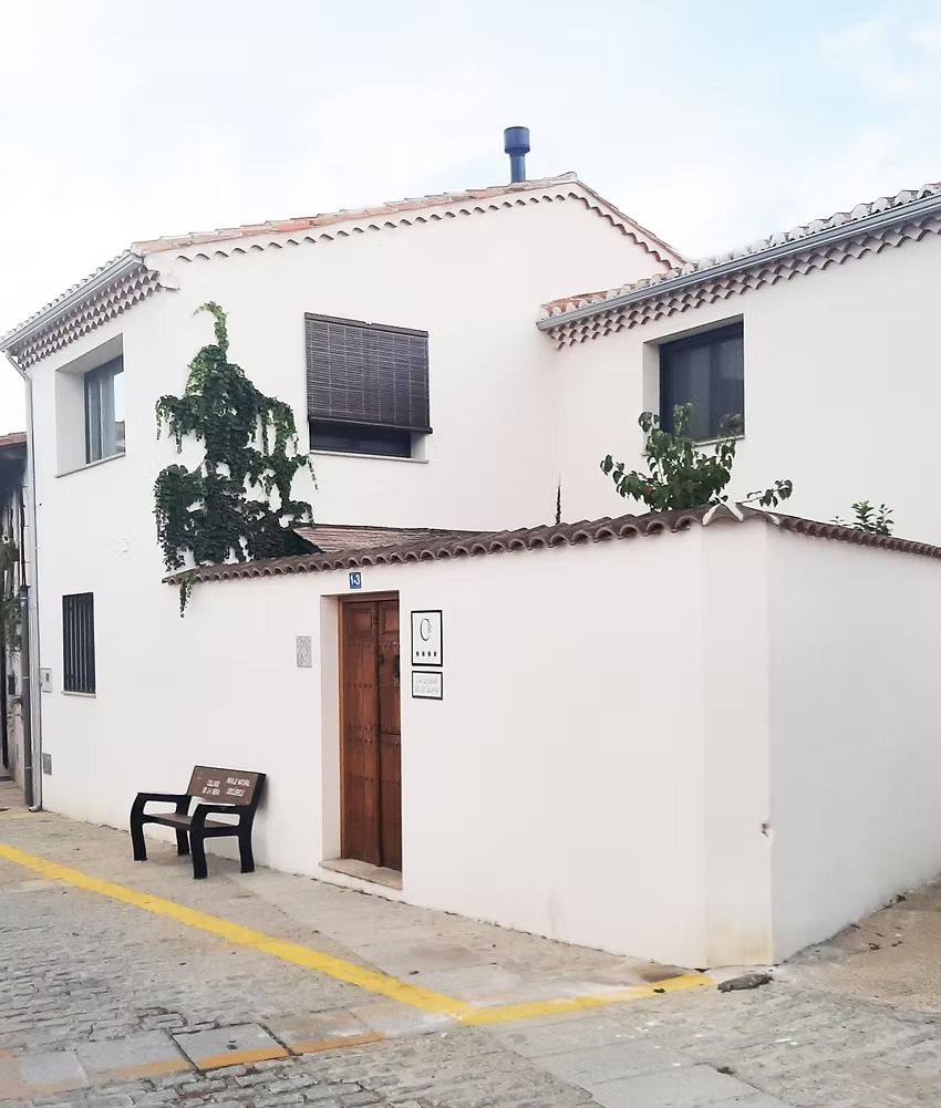 Casa blanca en una esquina de una calle empedrada, con puerta de madera, banco negro y plantas trepadoras.