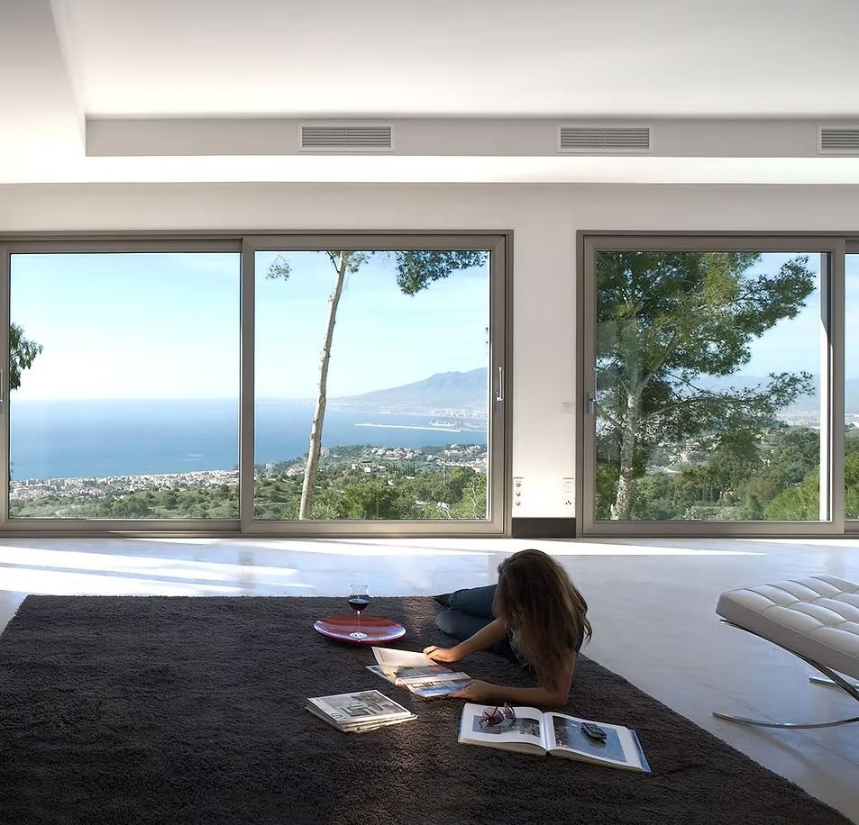 Una persona lee sobre una alfombra en una habitación moderna y luminosa con grandes ventanales con vistas al océano.