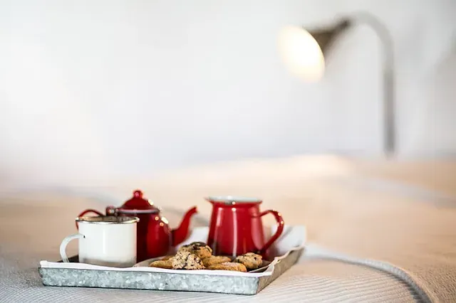 Bandeja con tetera roja, taza y galletas sobre una cama, debajo de una lámpara.