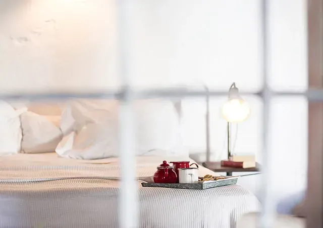 Una cama con sábanas blancas, una bandeja con un juego de té rojo y una lámpara encendida, vistas a través del marco de una ventana.