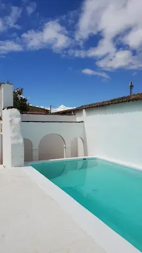 Un patio de paredes blancas con una piscina verde azulado bajo un cielo azul con nubes.