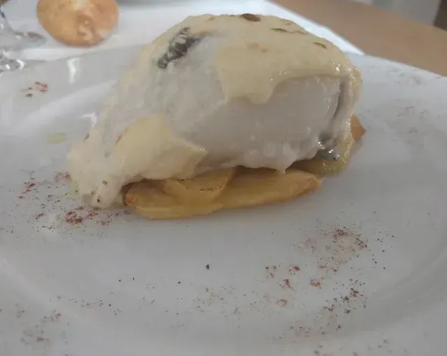 Pescado blanco cubierto con salsa, servido sobre una cama de patatas en un plato blanco.