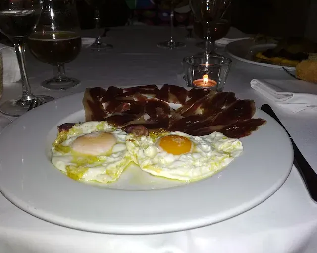 Huevos fritos y jamón curado en un plato blanco en una mesa, con vela encendida y bebidas.