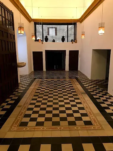 Pasillo con suelo a cuadros, puertas de madera oscura y lámparas colgantes, con luz que fluye desde una fuente invisible arriba.