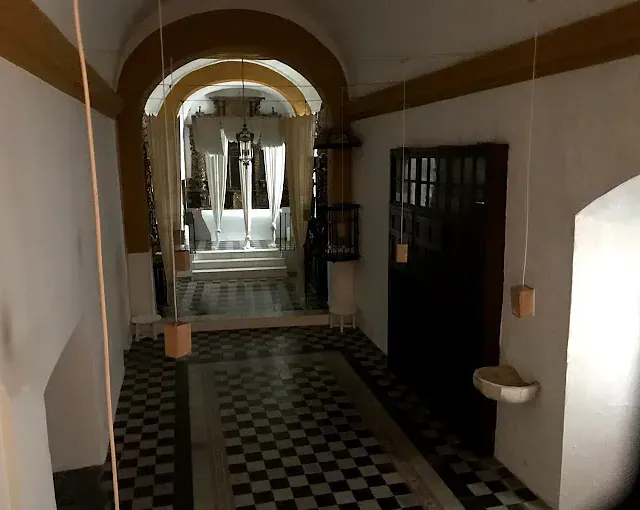 Pasillo con suelo cuadriculado, puerta arqueada a una habitación luminosa, cortinas blancas y puerta de madera oscura.