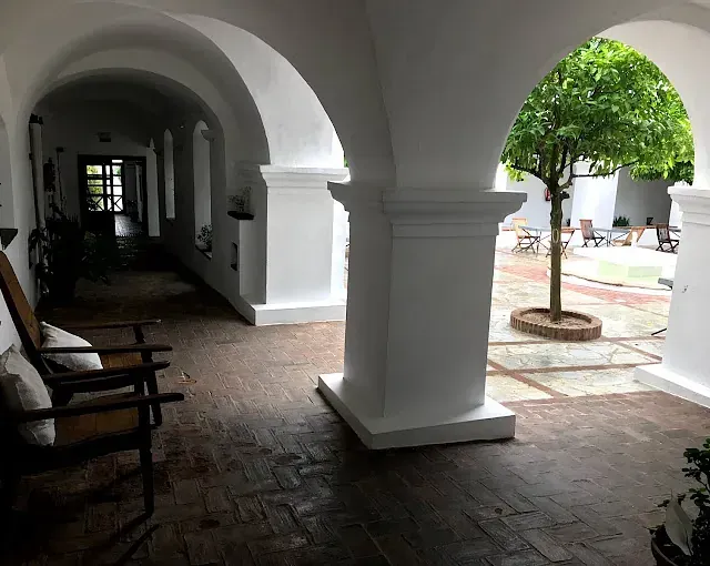 Pasarela cubierta con arcos blancos, que conduce a un patio con árbol, sillas y piso de ladrillo.