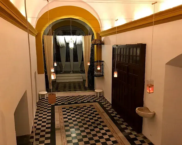 Interior con suelo de tablero de ajedrez, entrada arqueada, molduras amarillas y faroles colgantes.