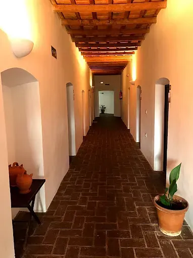 Largo pasillo con puertas arqueadas, suelo de ladrillo y techo de madera. La luz del sol proyecta sombras.