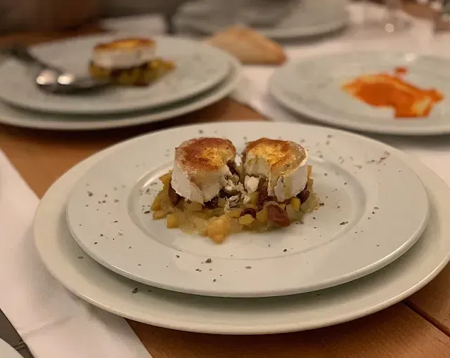 Medallones de queso de cabra sobre cama de patatas y nueces, espolvoreados con condimentos, servidos en un plato blanco.