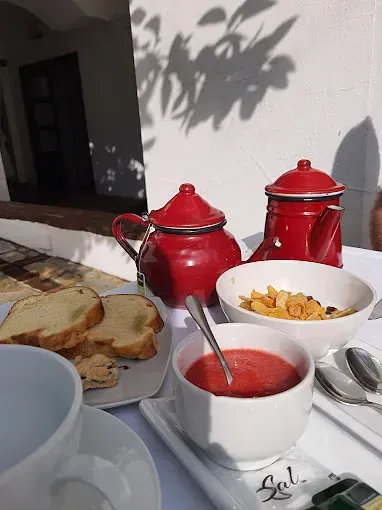 Desayuno servido sobre una superficie blanca: teteras rojas, pan, cereales, sopa de tomate y una taza de té en un día soleado.