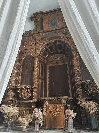 Altar ornamentado, de color dorado, en el interior, flanqueado por cortinas blancas y jarrones con flores secas.