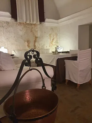 Una olla de cobre cuelga en una habitación rústica. Sábanas blancas sobre la cama y mesa preparada para comer.