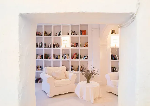 Habitación blanca con estanterías empotradas, sillones y una pequeña mesa con flores.