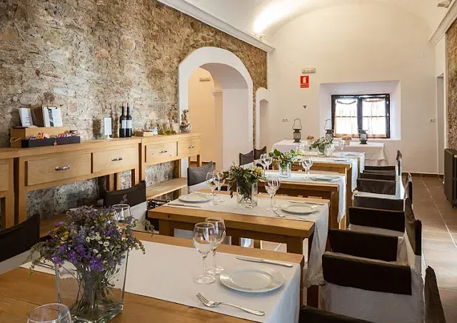 Interior de restaurante con largas mesas de madera dispuestas para los comensales, pared de piedra, mantelería blanca y centros de mesa de flores.