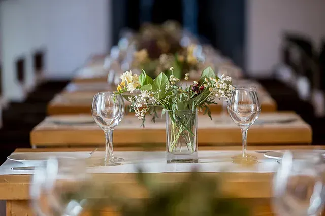 Largas mesas de madera preparadas para comer, con cristalería, platos y centros de mesa con flores.