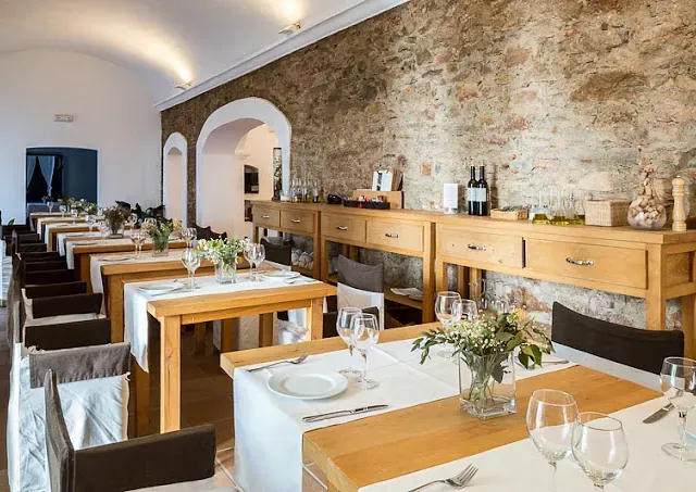 Interior del restaurante: mesas de madera con manteles blancos, pared de piedra, puertas arqueadas, preparadas para los comensales.