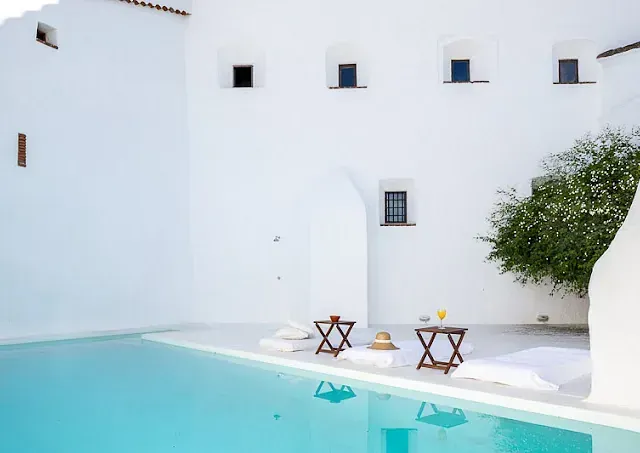 Patio de paredes blancas con piscina, dos mesas pequeñas y un árbol en flor.