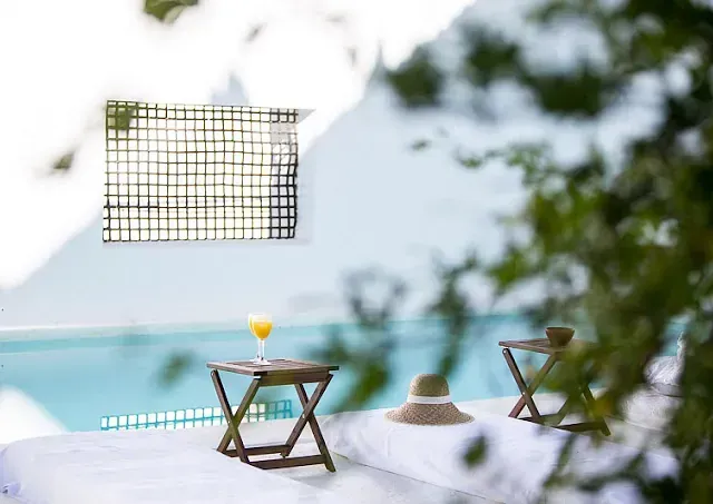 Escena junto a la piscina: cóctel en una mesa, sombrero y dos mesas plegables de madera cerca de la piscina. Luz solar brillante, follaje borroso.