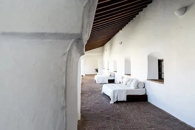 Largo pasillo blanco con camas, ventanas arqueadas y un techo de madera oscura.