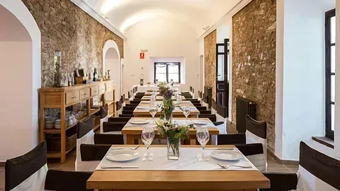 Comedor largo con largas mesas de madera preparadas para comer, paredes de piedra y centros de mesa florales.