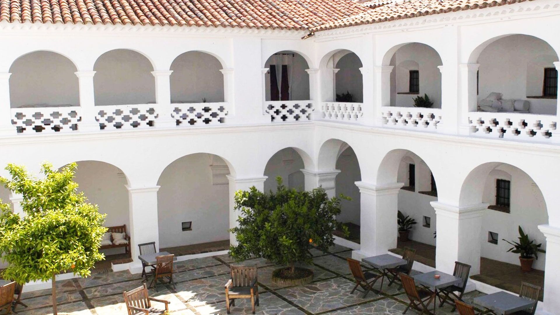 Patio blanco con pasillos arqueados, mesas, sillas y vegetación; techo de tejas rojas en lo alto.
