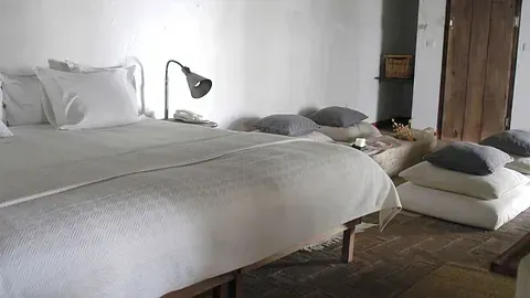 Dormitorio con paredes blancas, cama grande, cojines en el suelo y puerta de madera.