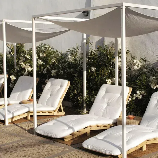 Cuatro sillones blancos con toldos colocados en un patio de piedra, rodeados de vegetación y una pared blanca.