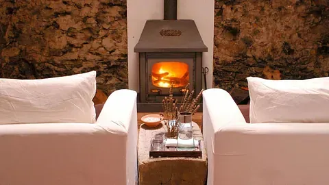 Dos sillones blancos frente a una chimenea con fuego ardiente en una acogedora habitación con paredes de piedra.