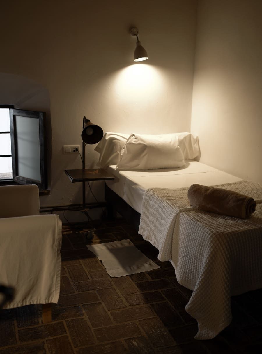 Dormitorio con cama individual, lámpara de lectura, sábanas blancas y suelo de ladrillo. Iluminación tenue, ventana visible.