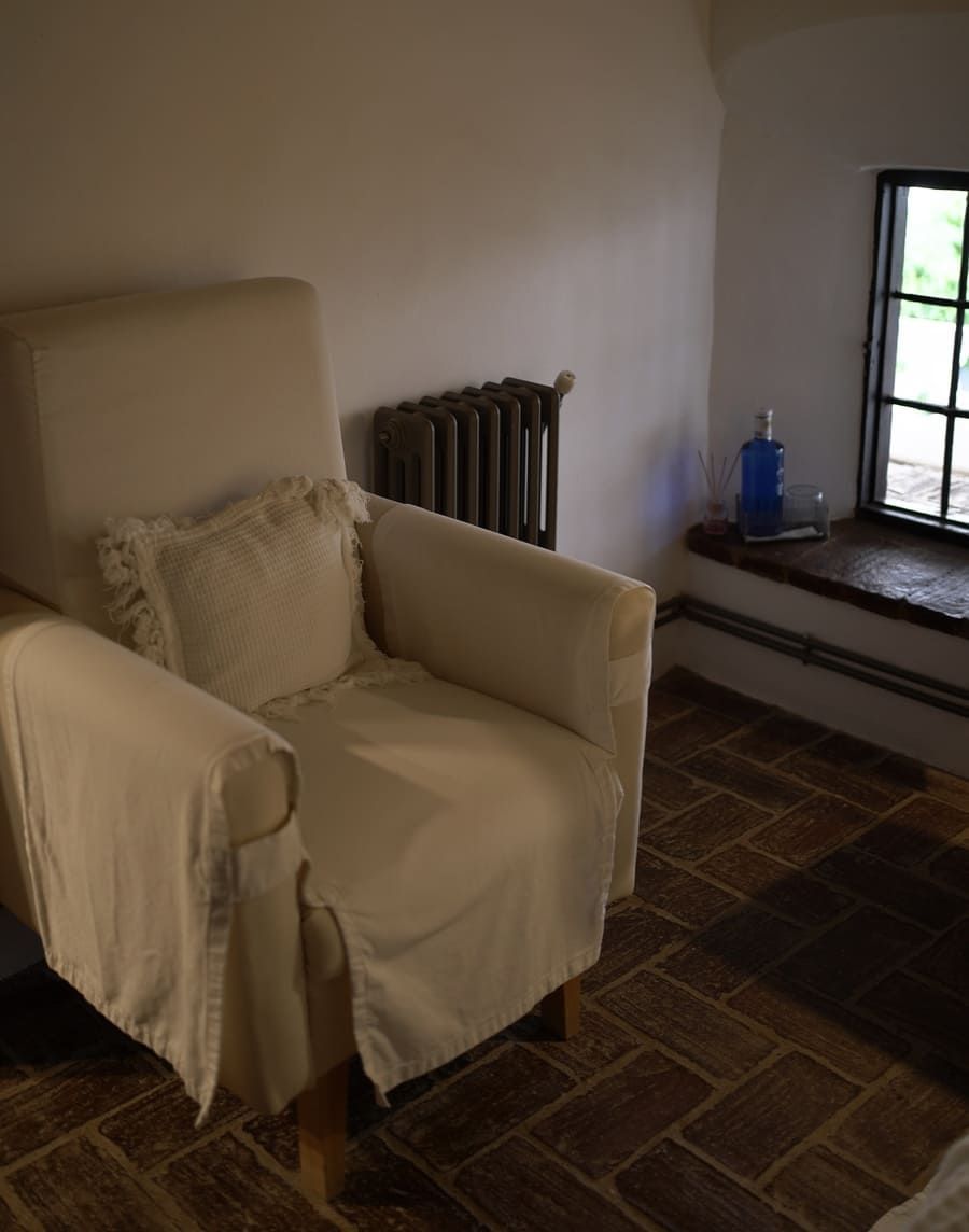 Sillón color crema en una habitación con ventana, radiador y suelo de ladrillo.