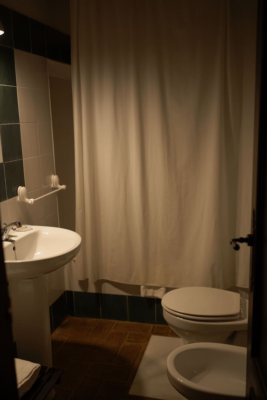 Baño con lavabo, inodoro y cortina de ducha blancos. Azulejos verdes y suelo marrón.