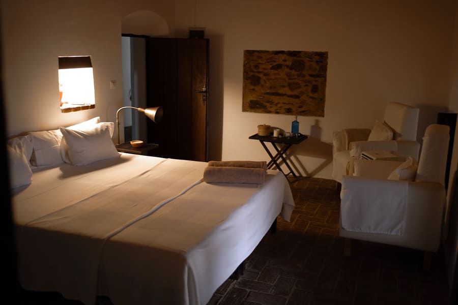 Dormitorio con cama tendida, mesita de noche, sillón y cuadros en la pared. Luz tenue, colores blanco y tostado.