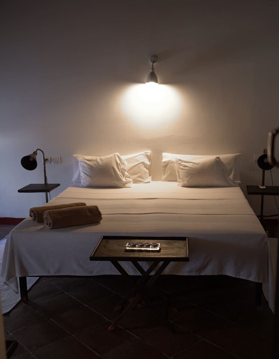 Dormitorio con ropa de cama blanca, dos almohadas, una mesita de noche oscura con una lámpara a cada lado, una bandeja con un control remoto sobre la cama.